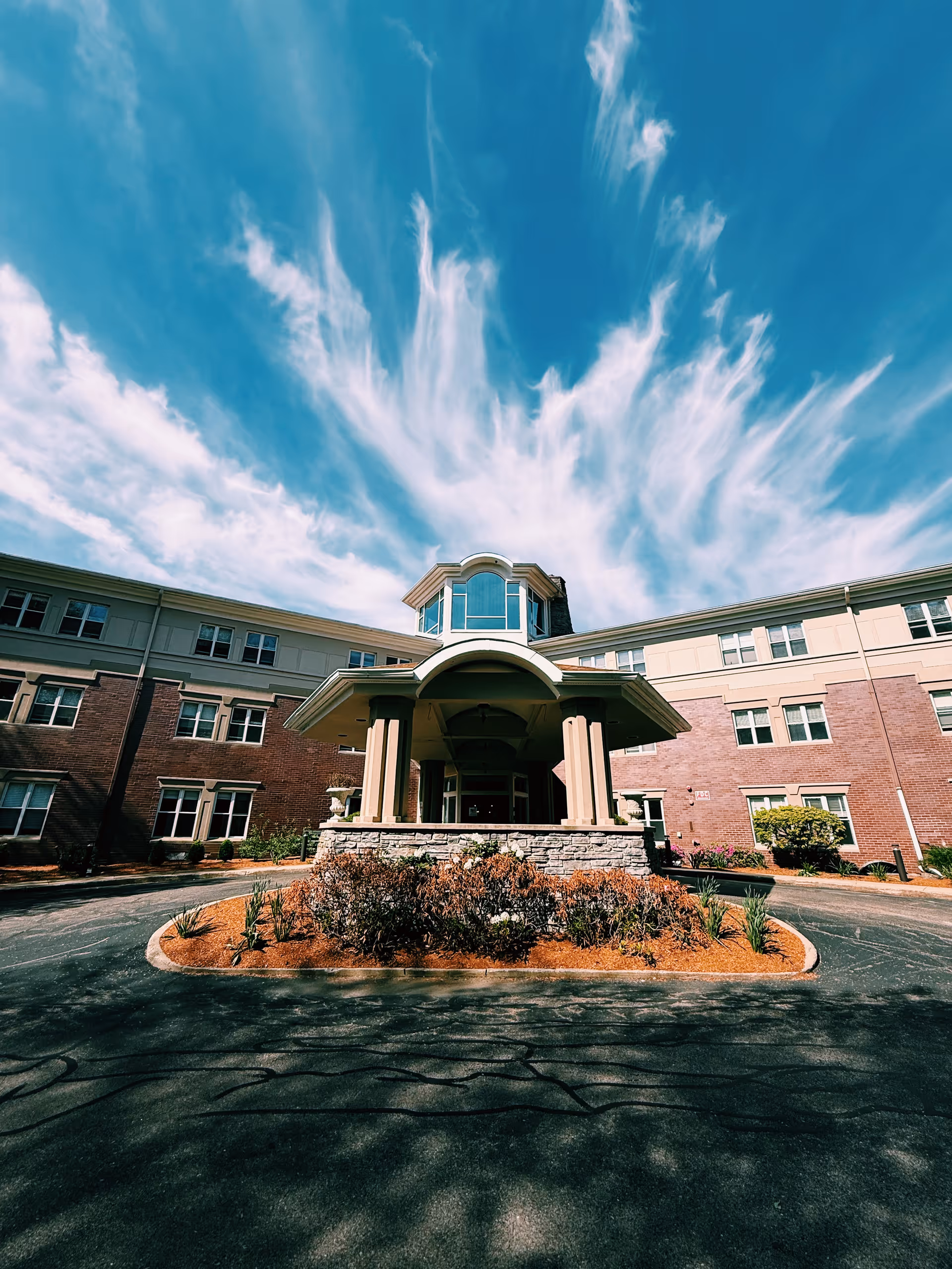Front entrance of a multi-story brick assisted living building with a covered porte-cochere and circular driveway under a blue sky.