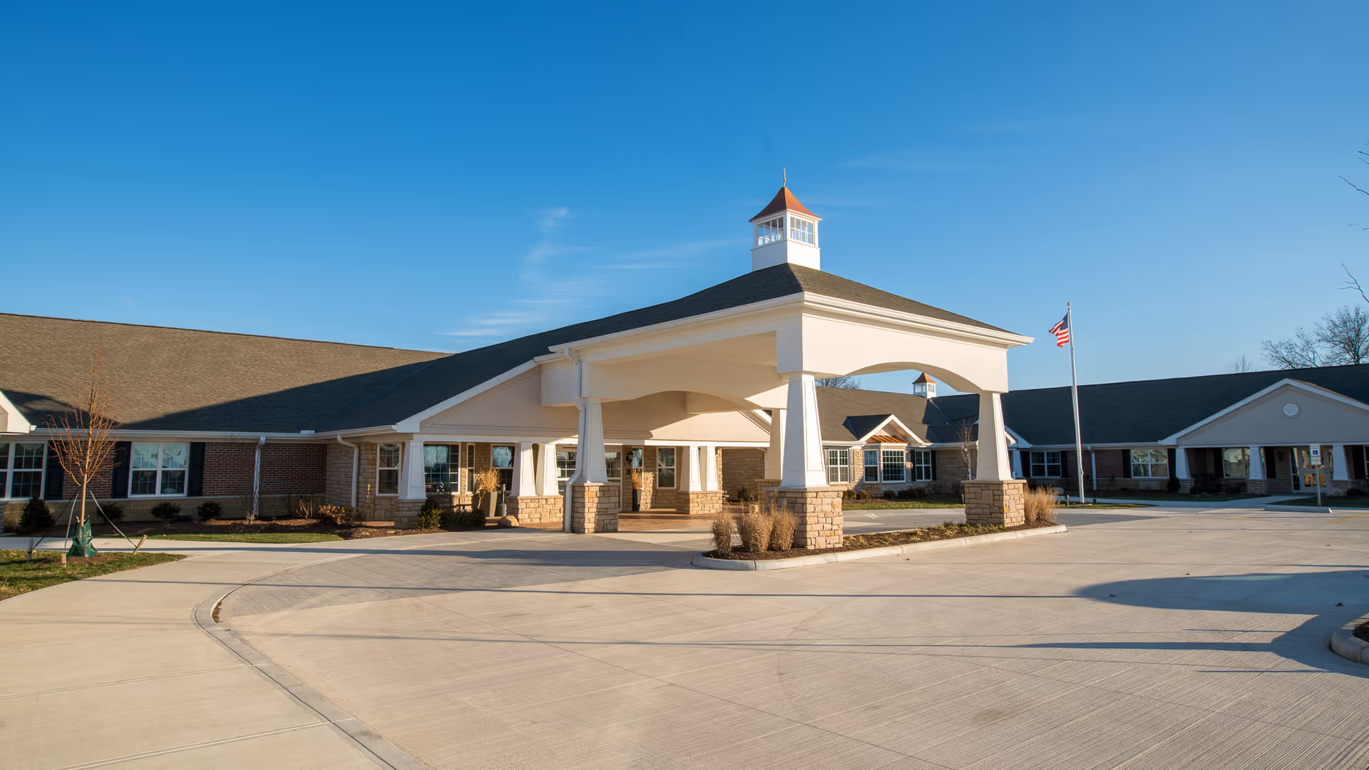 Exterior view of Capri Gardens Nursing Center showing a large covered entrance with white pillars and a cupola on the roof. The building is single-story with brick and stone accents, surrounded by a paved driveway and an American flag on a flagpole. The sky is clear and blue.