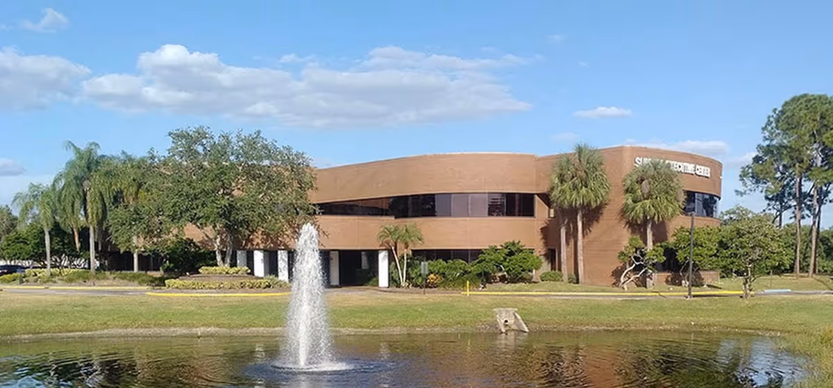 Exterior view of a brown curved senior living facility with a fountain pond and palm trees.