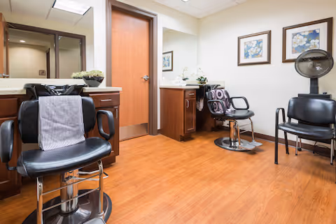 Interior view of a hair salon area in a senior living facility with wooden flooring, two black salon chairs, a hair dryer, a counter with a mirror, and framed floral artwork on the wall.
