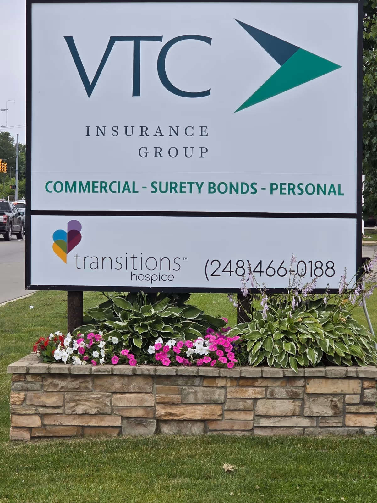 Outdoor sign for VTC Insurance Group and Transitions Hospice beside a flower bed.