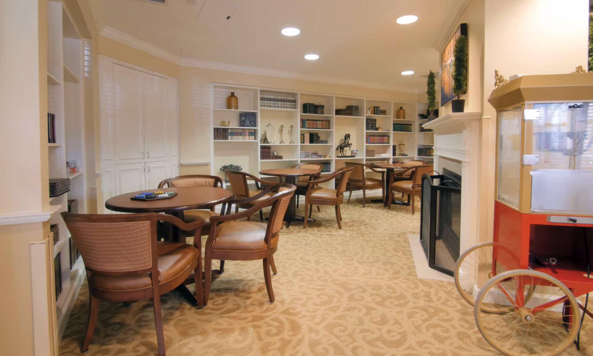 Spacious indoor senior living lounge with round tables, comfortable chairs, bookshelves, and a fireplace.