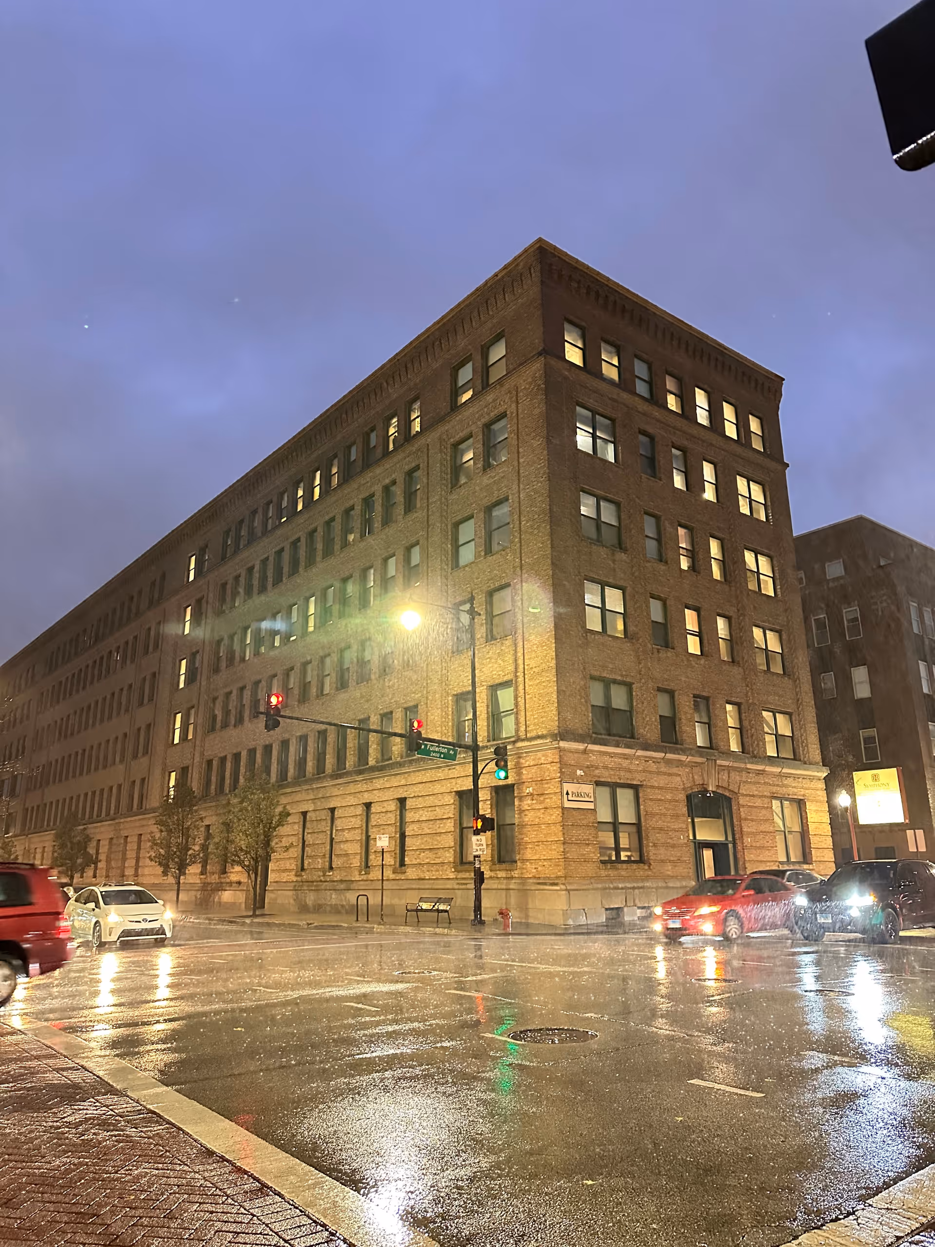 A large multi-story brick building at a street corner during rainy evening. Several cars are stopped at a traffic light with wet streets reflecting the lights. The building has many windows, some illuminated, and a street sign reading Fullerton Ave is visible.