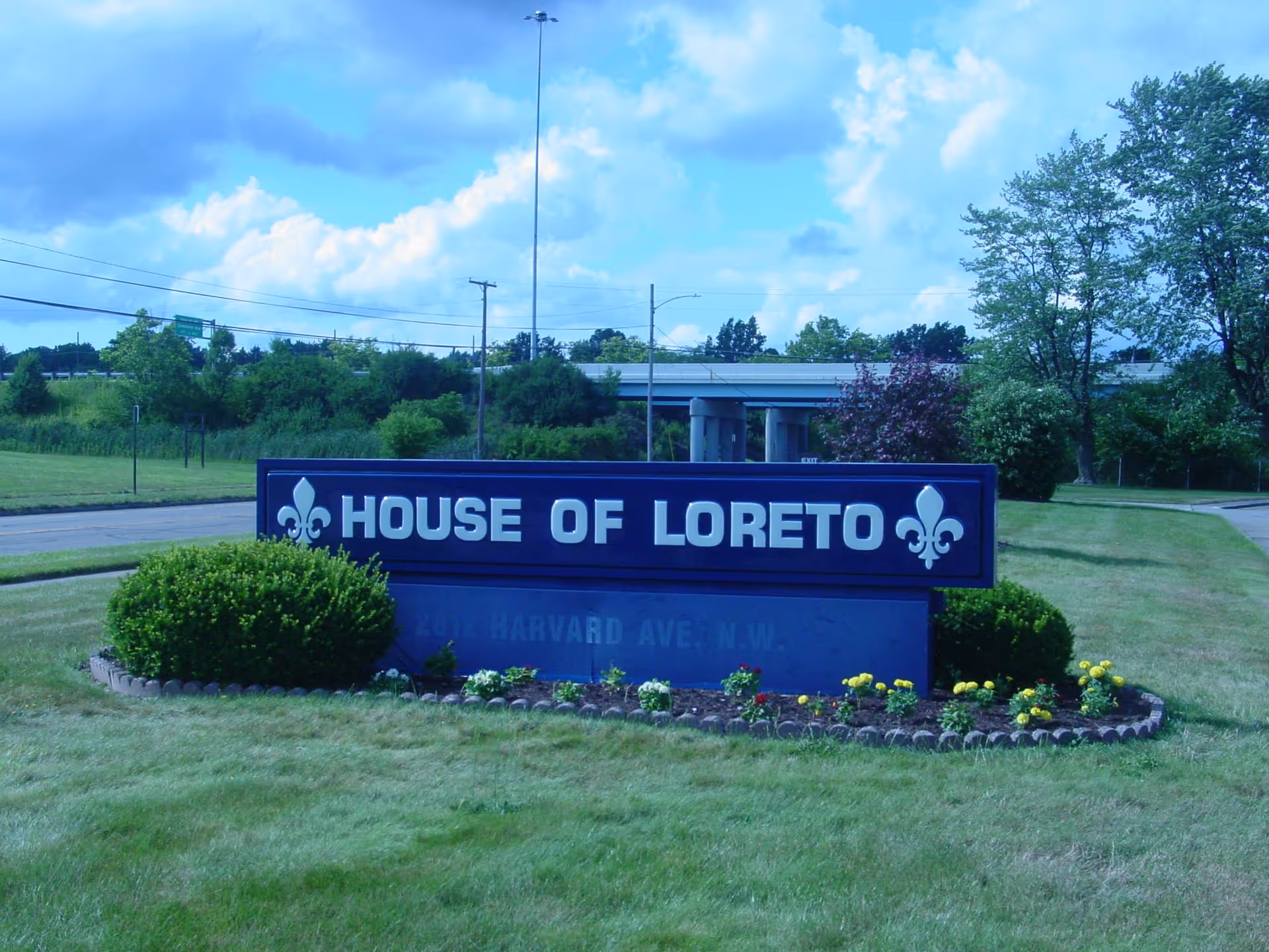 Blue House of Loreto sign on a grassy lawn with shrubs and trees.