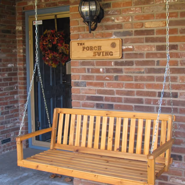 A wooden porch swing hanging by metal chains on a brick wall porch. Above the swing is a wooden sign that reads 'The Porch Swing' and a black outdoor wall lantern. A door with a fall-themed wreath is visible in the background.