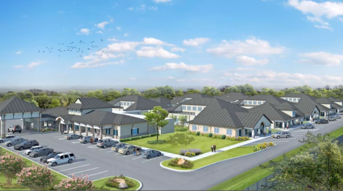 Aerial view of Greenbriar Community Care Center showing multiple connected buildings with gray roofs, surrounded by parking lots and green landscaped areas under a partly cloudy blue sky.
