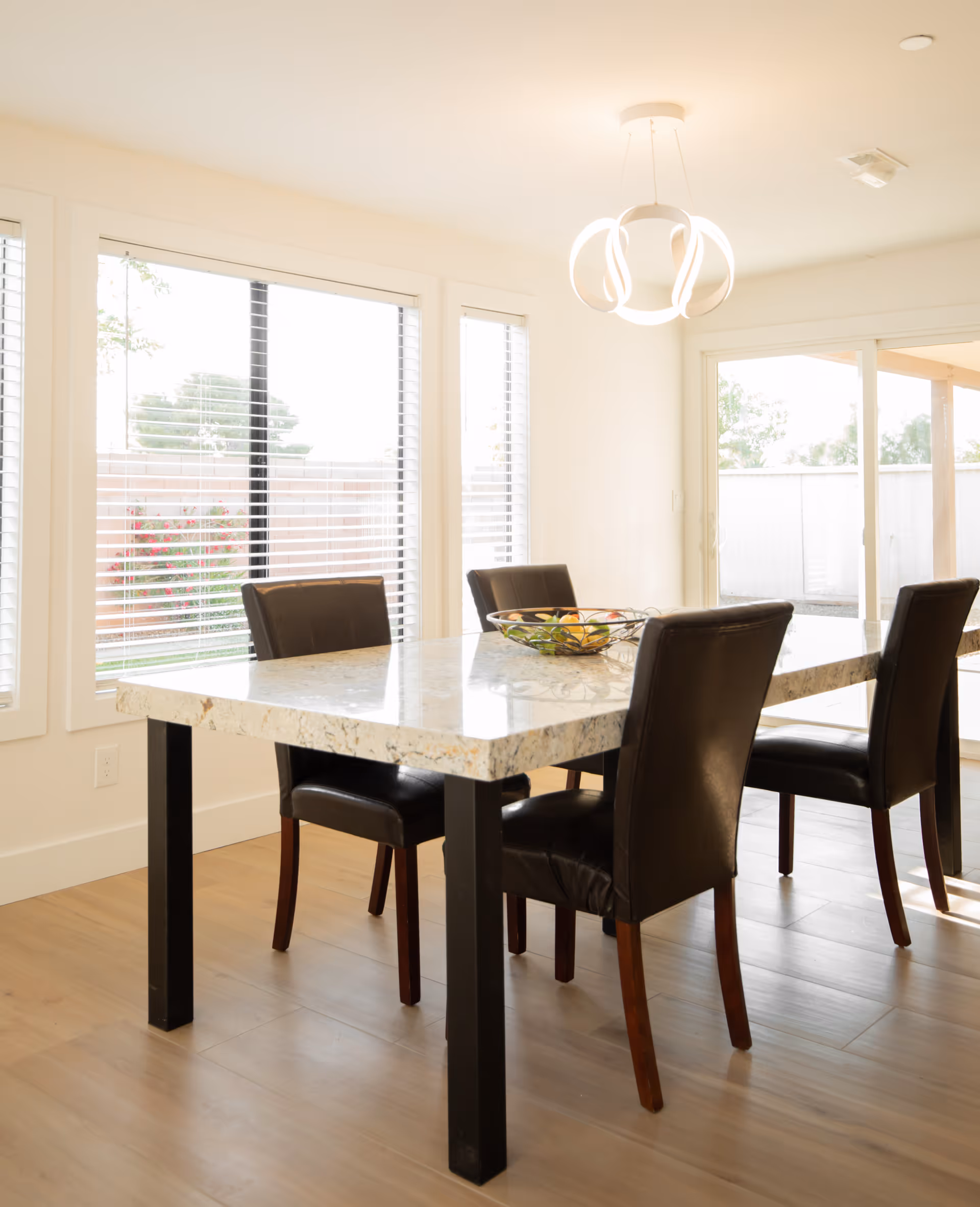 A bright dining area with a rectangular marble table and four dark brown leather chairs. Large windows with white blinds allow natural light to fill the room. A modern circular light fixture hangs above the table, and a bowl of fruit is placed on the table. Sliding glass doors lead to an outdoor space.