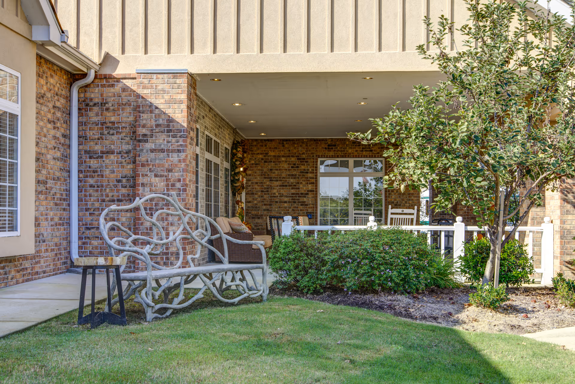 Covered brick front porch with decorative bench, rocking chairs, and landscaped lawn at an assisted living facility.