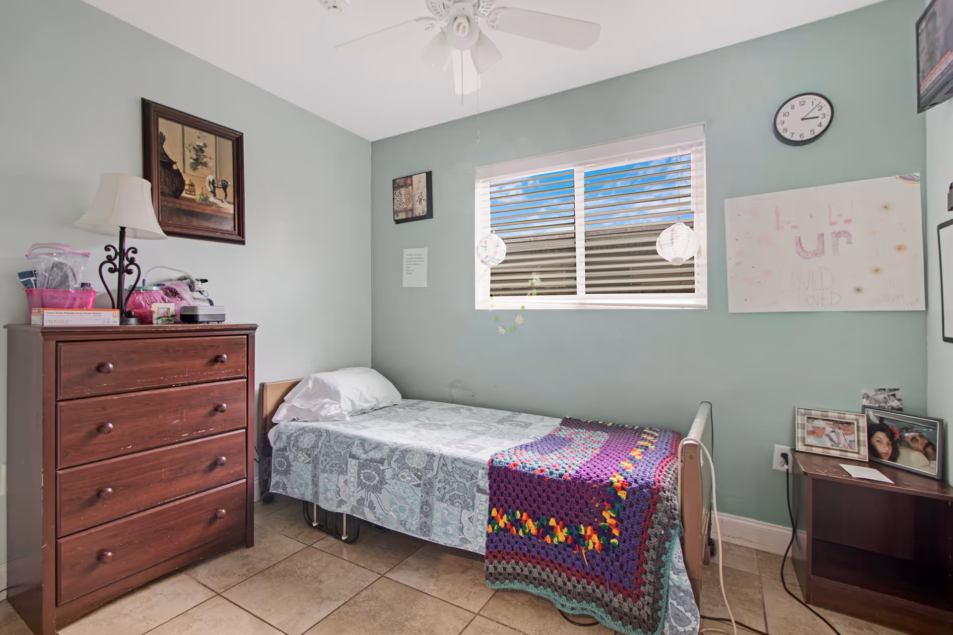 A small bedroom with a single bed, dresser, window, wall clock, and crocheted blanket.