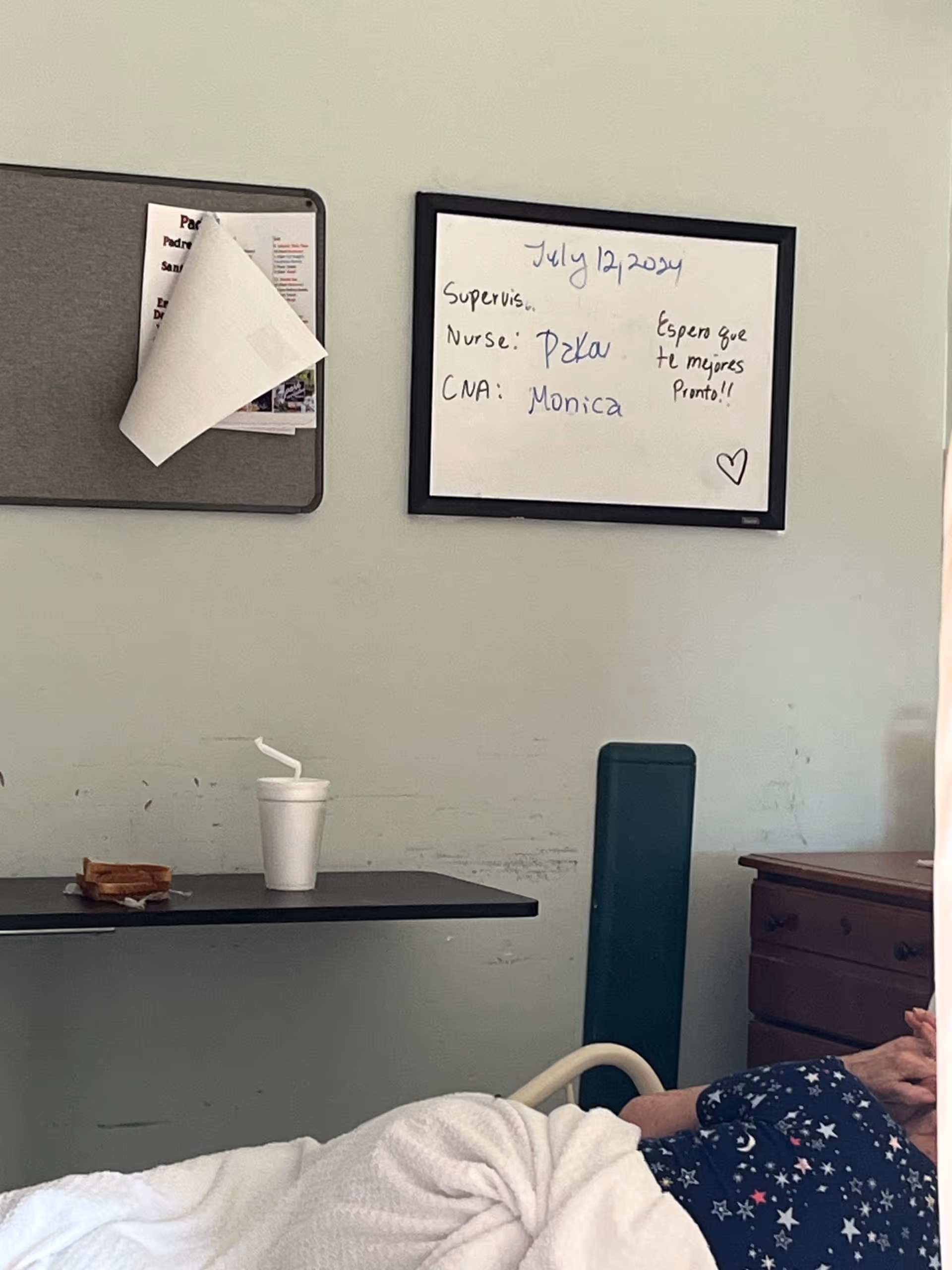 A pale-walled interior patient room showing bed, small table, whiteboard, and bulletin board.