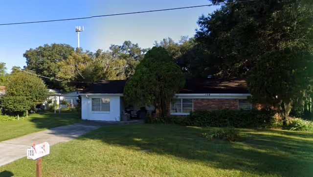 Single-story brick and siding residential building with a front lawn, driveway, mailbox, and trees.