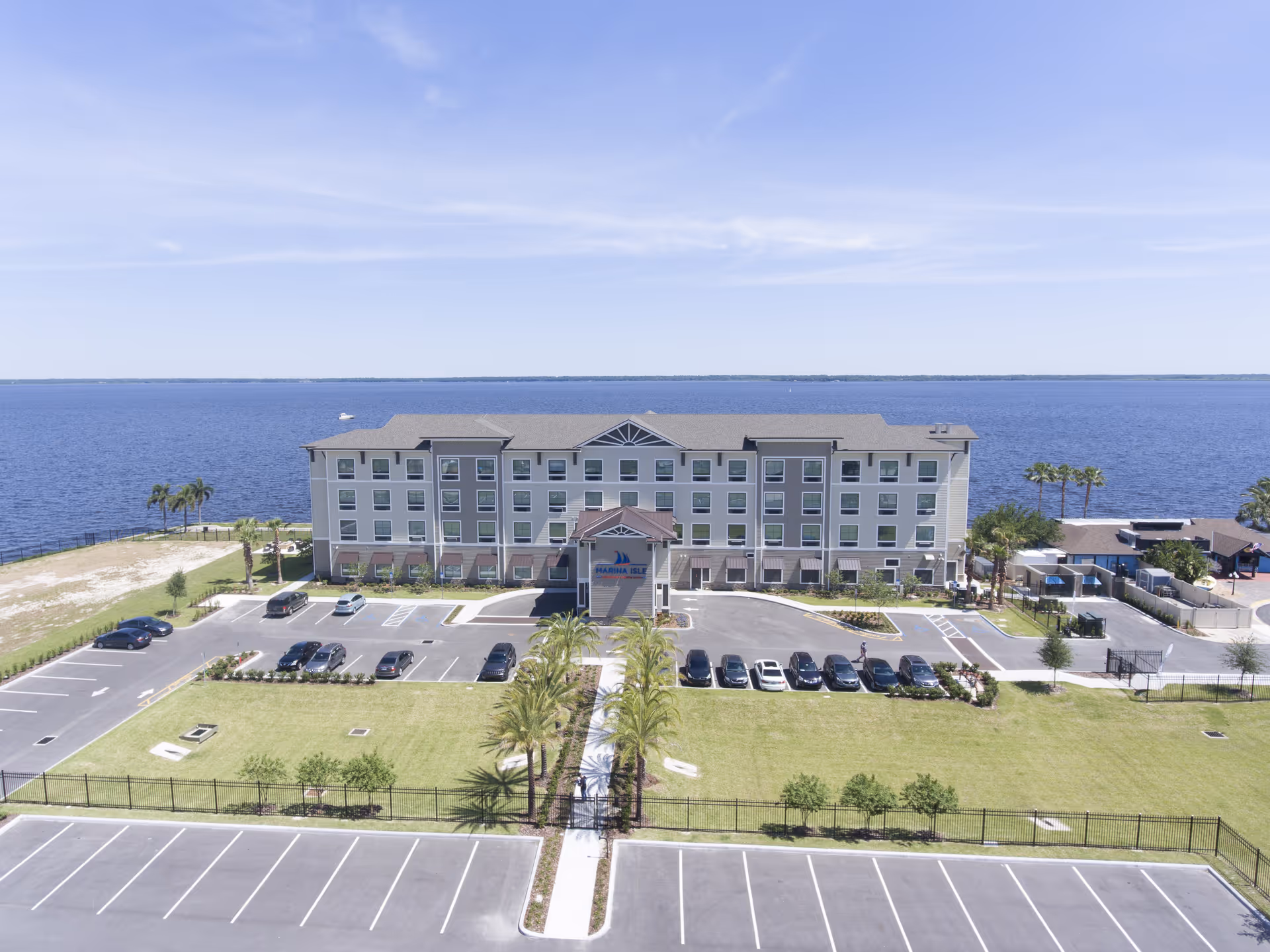 Four-story waterfront assisted living building facing a large body of water with a parking lot, lawn, and palm trees.