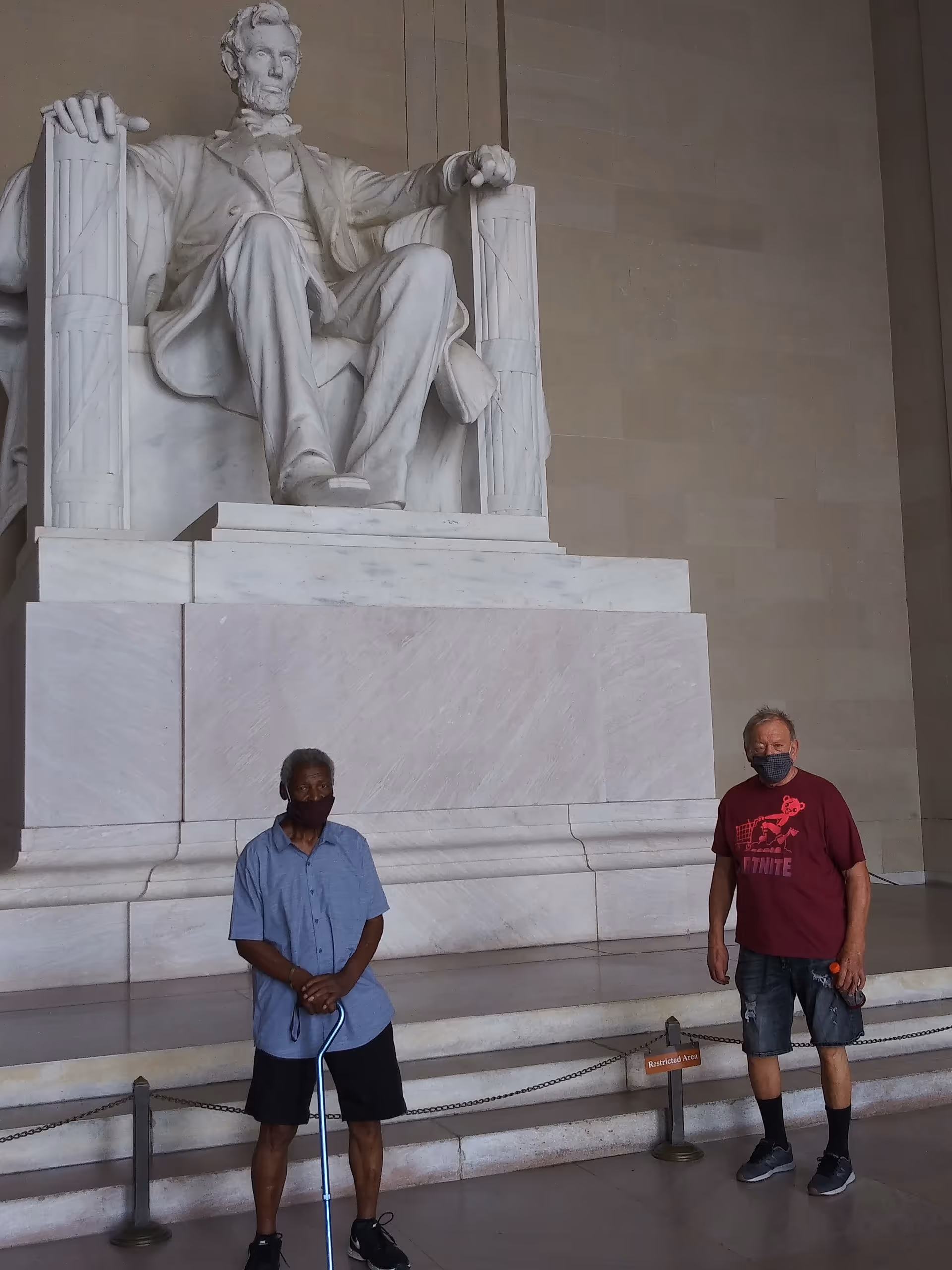 Two masked individuals stand near a large seated marble statue indoors.