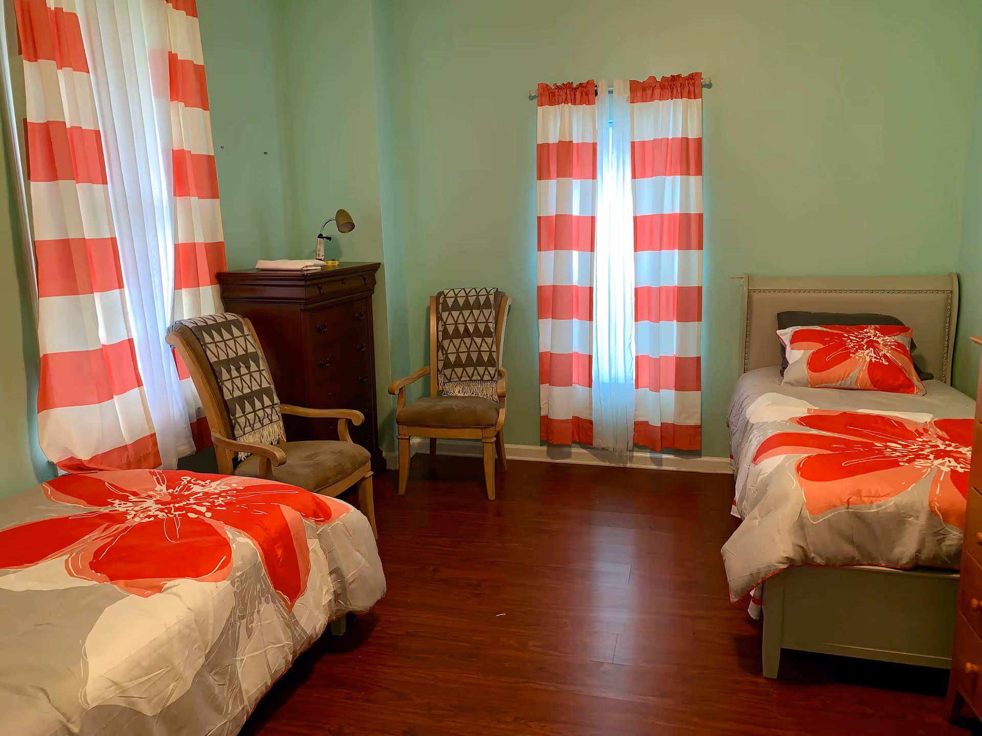 Two-bed bedroom with teal walls, red-and-white striped curtains, matching floral bedding, two armchairs and a wooden dresser.