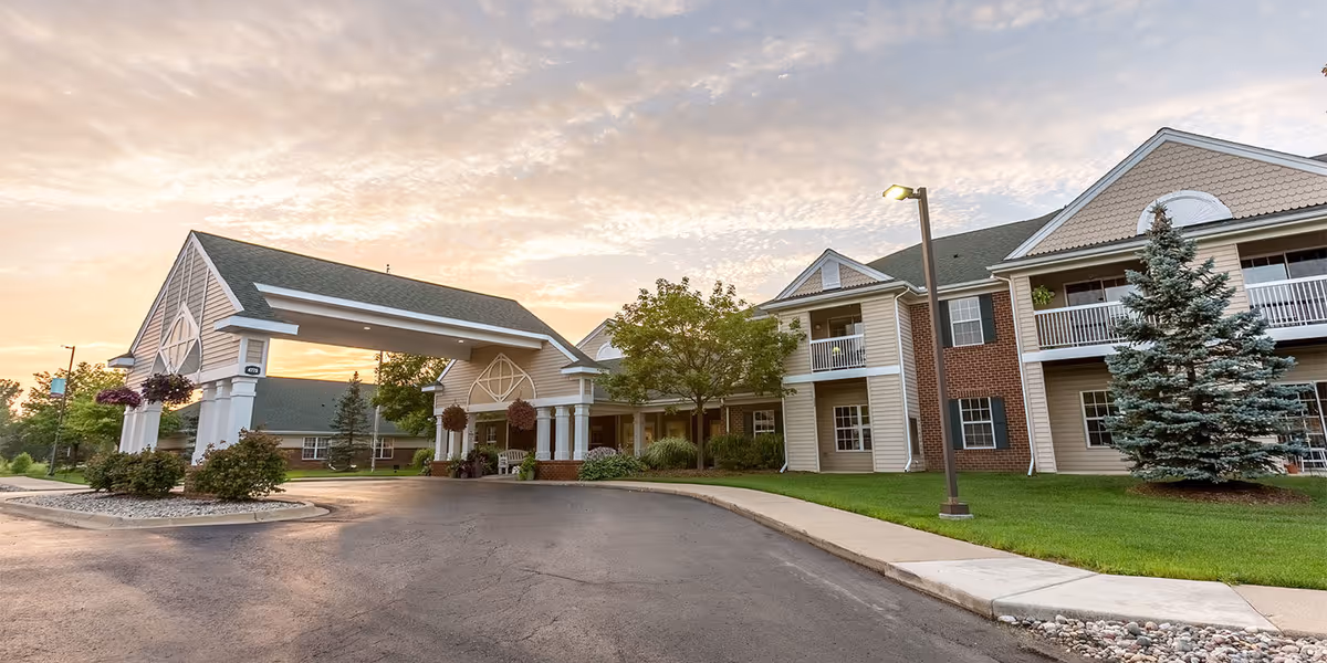 Exterior view of Independence Village of Grand Ledge senior living facility at sunset, showing a covered entrance with columns, well-maintained landscaping including trees and shrubs, and a two-story building with balconies and windows.