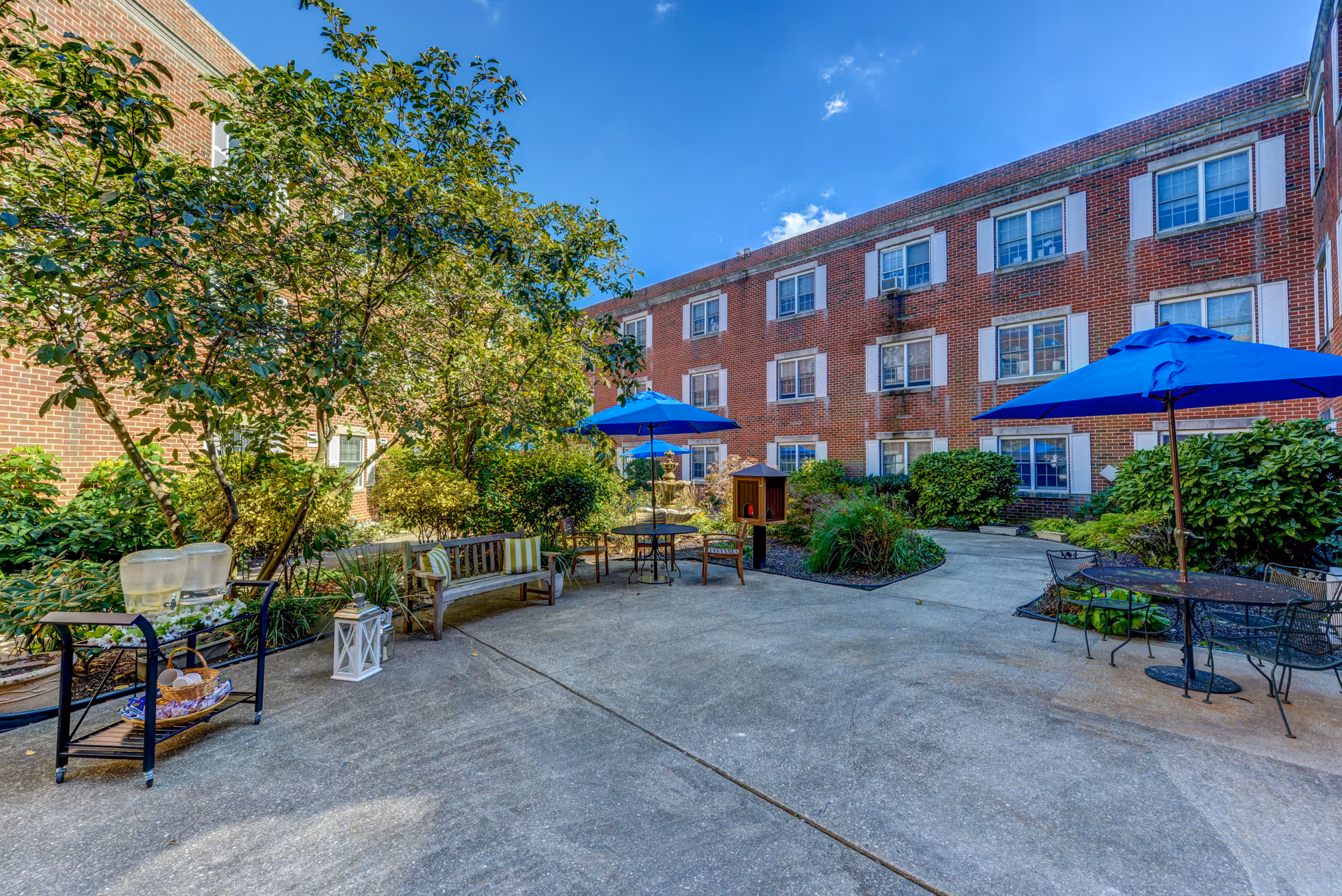 Sunlit courtyard with patio tables, blue umbrellas, benches and landscaping surrounded by a three-story brick building.