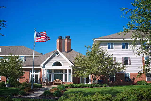 Two-story senior living facility with brick, white siding, flagpole, and landscaping.