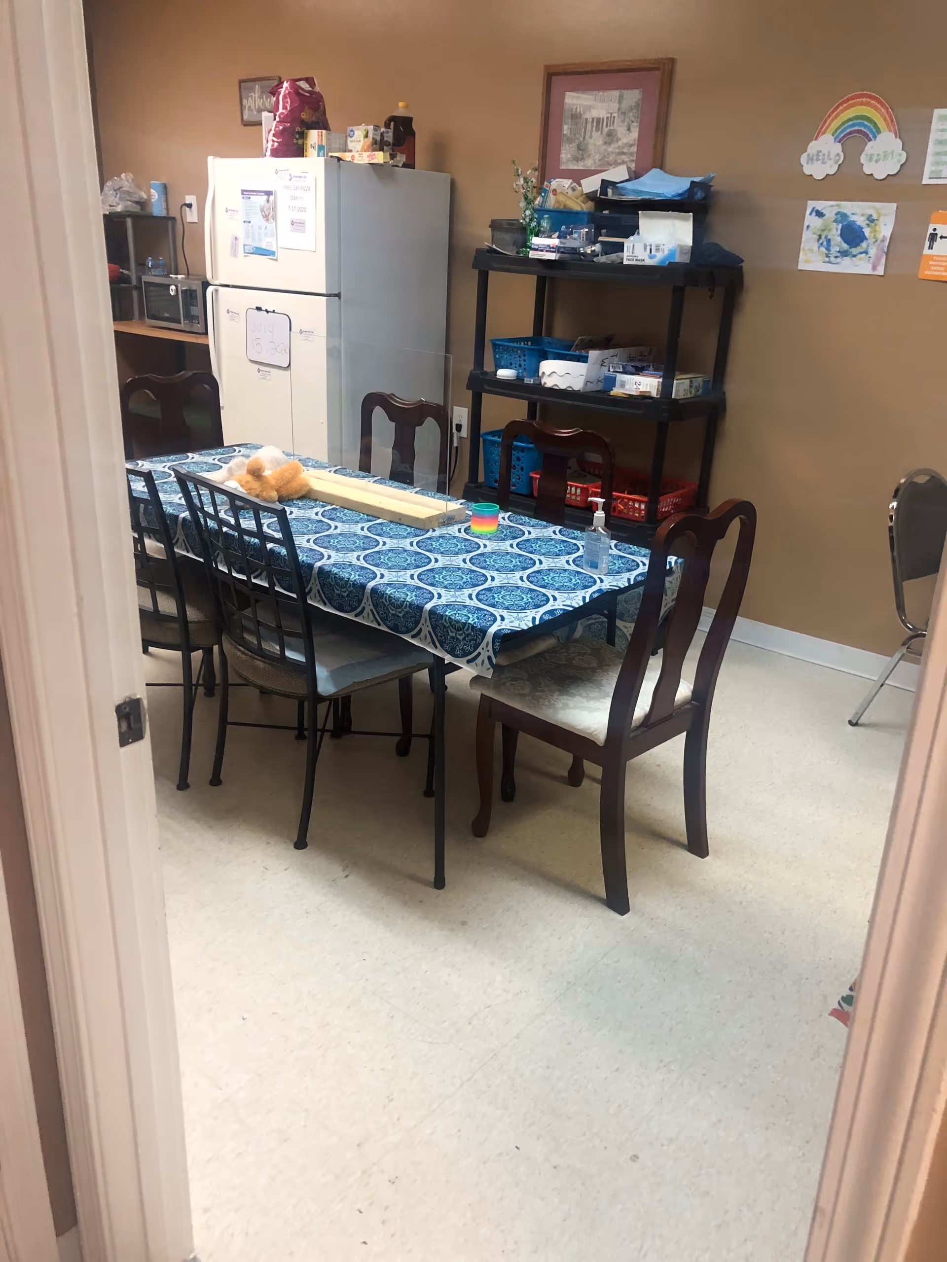 A small room with a table covered by a blue patterned tablecloth surrounded by six chairs. On the table are a teddy bear, a clear plastic divider, a colorful toy, and a bottle of hand sanitizer. In the background, there is a white refrigerator with papers attached to it, a black shelving unit with various items, and children's artwork on the beige walls.