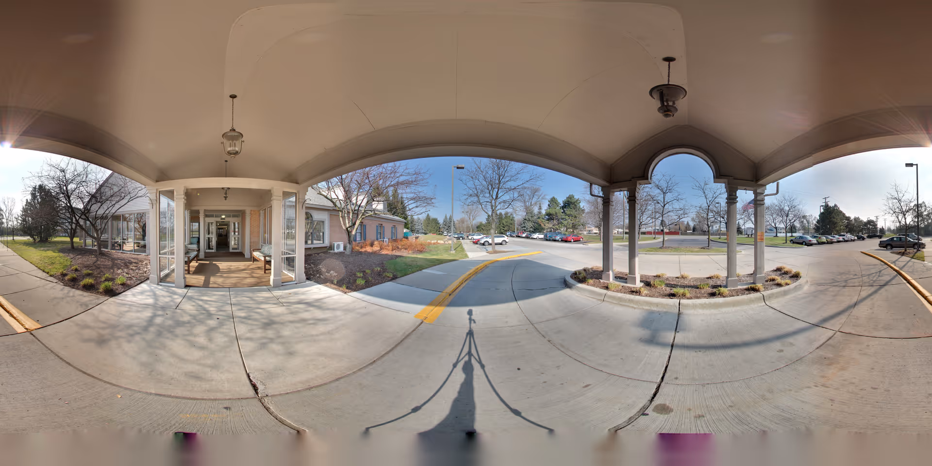 Covered entrance area of Oakmont Livonia Senior Community with a concrete driveway and sidewalk, landscaped garden beds with small shrubs, leafless trees, and a parking lot with several cars in the background under a clear sky.