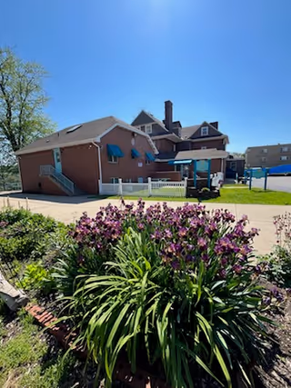 Exterior view of a brick building with blue awnings and a white fence, surrounded by a garden with purple flowers and green plants under a clear blue sky.