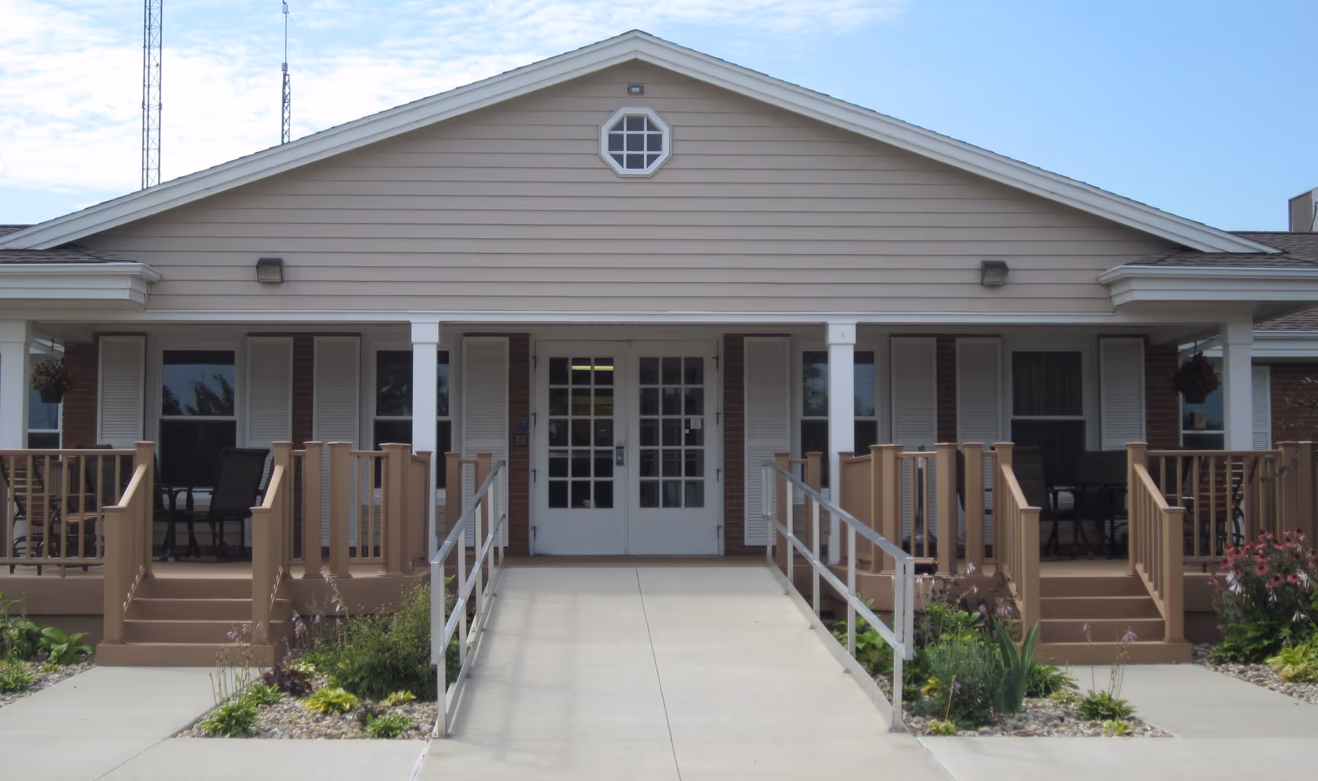 Front entrance of a single-story residential building with a wheelchair ramp, porches with chairs, and double glass doors under a gabled roof.