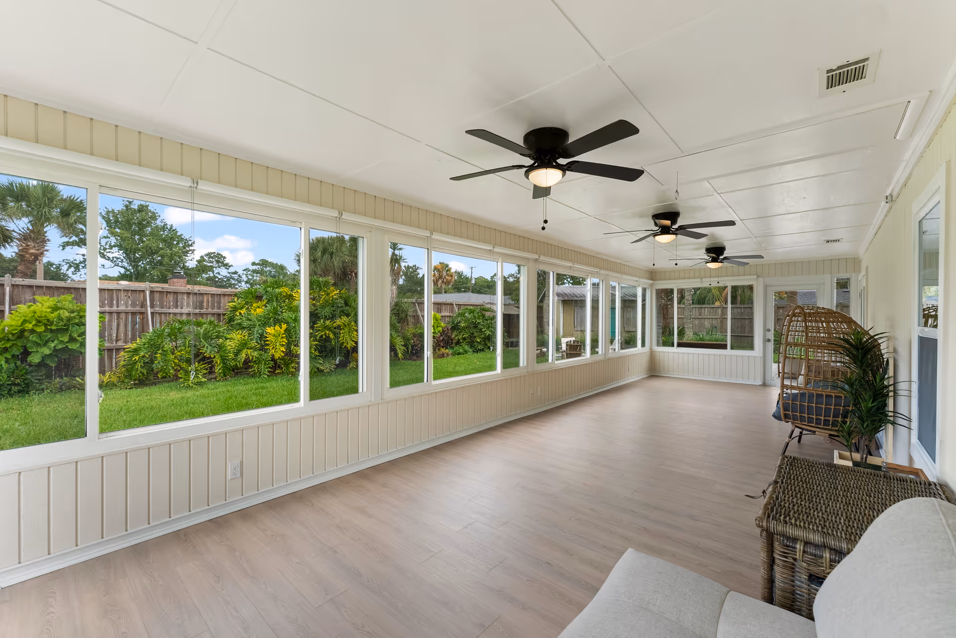 Bright enclosed sunroom with large windows, ceiling fans, wicker seating, and garden view.