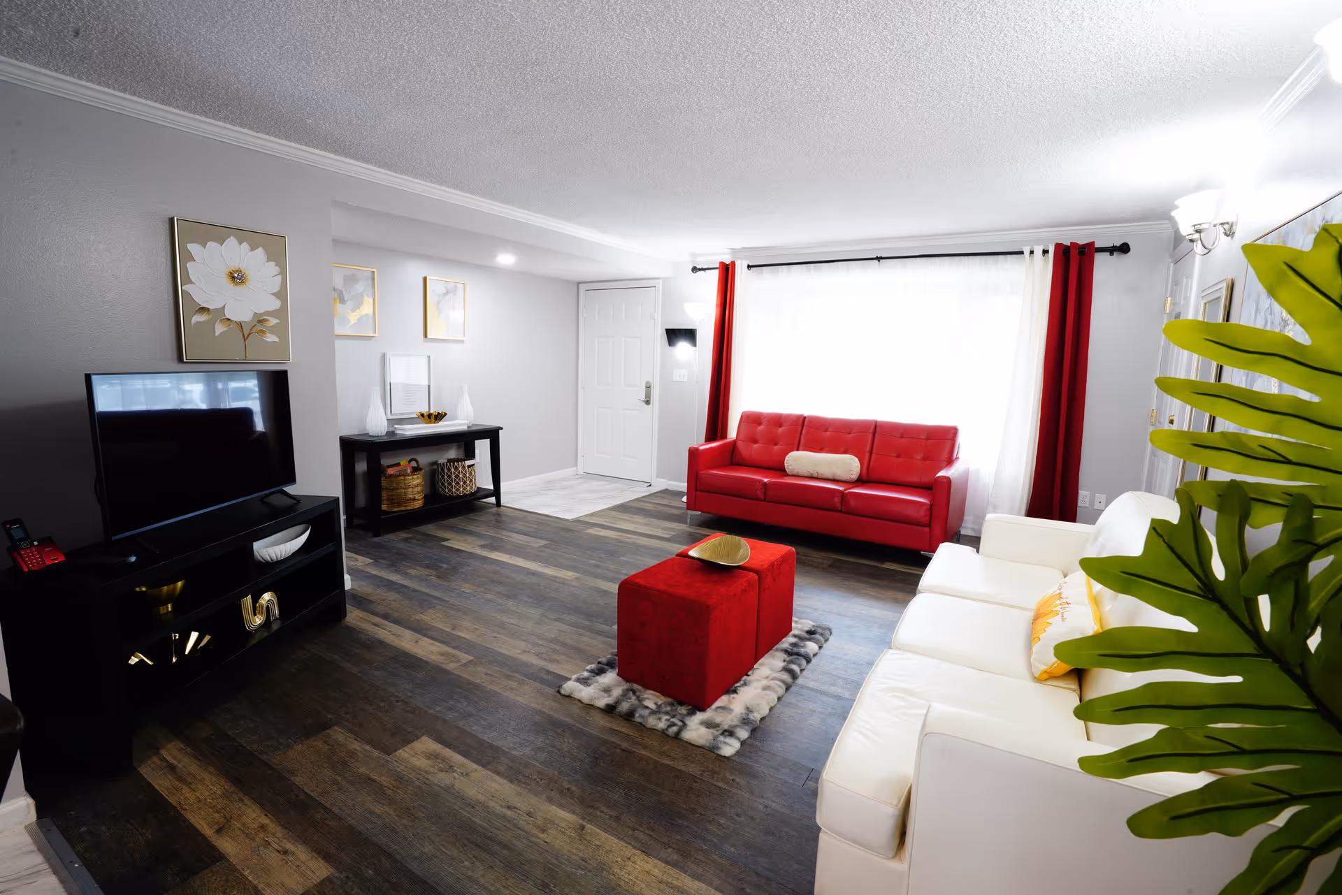 Bright living room with white couches, a red sofa, ottoman, TV, and plant.