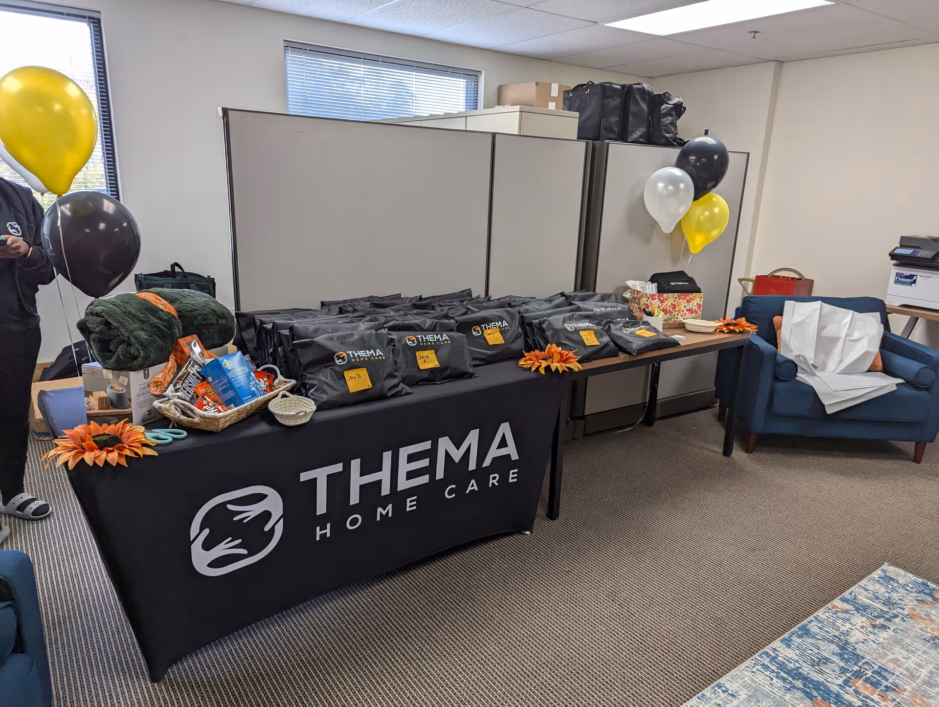A table with Thema Home Care gift bags and balloons in a small office room.
