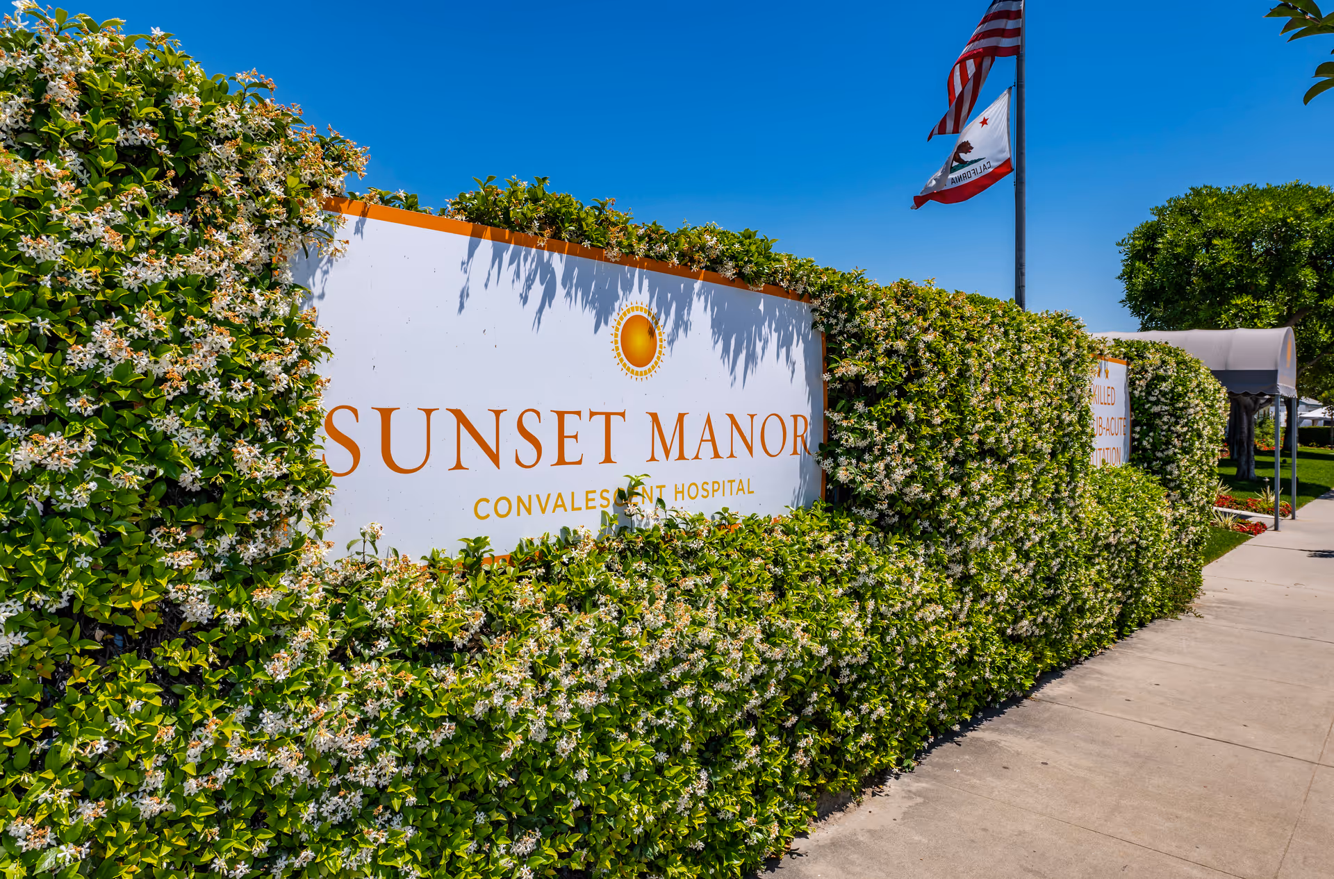 A large white sign with orange text reading 'Sunset Manor Convalescent Hospital' partially covered by green leafy bushes with white flowers. Two flags, one American and one California state flag, are flying on a flagpole behind the sign. A sidewalk runs alongside the bushes under a clear blue sky.