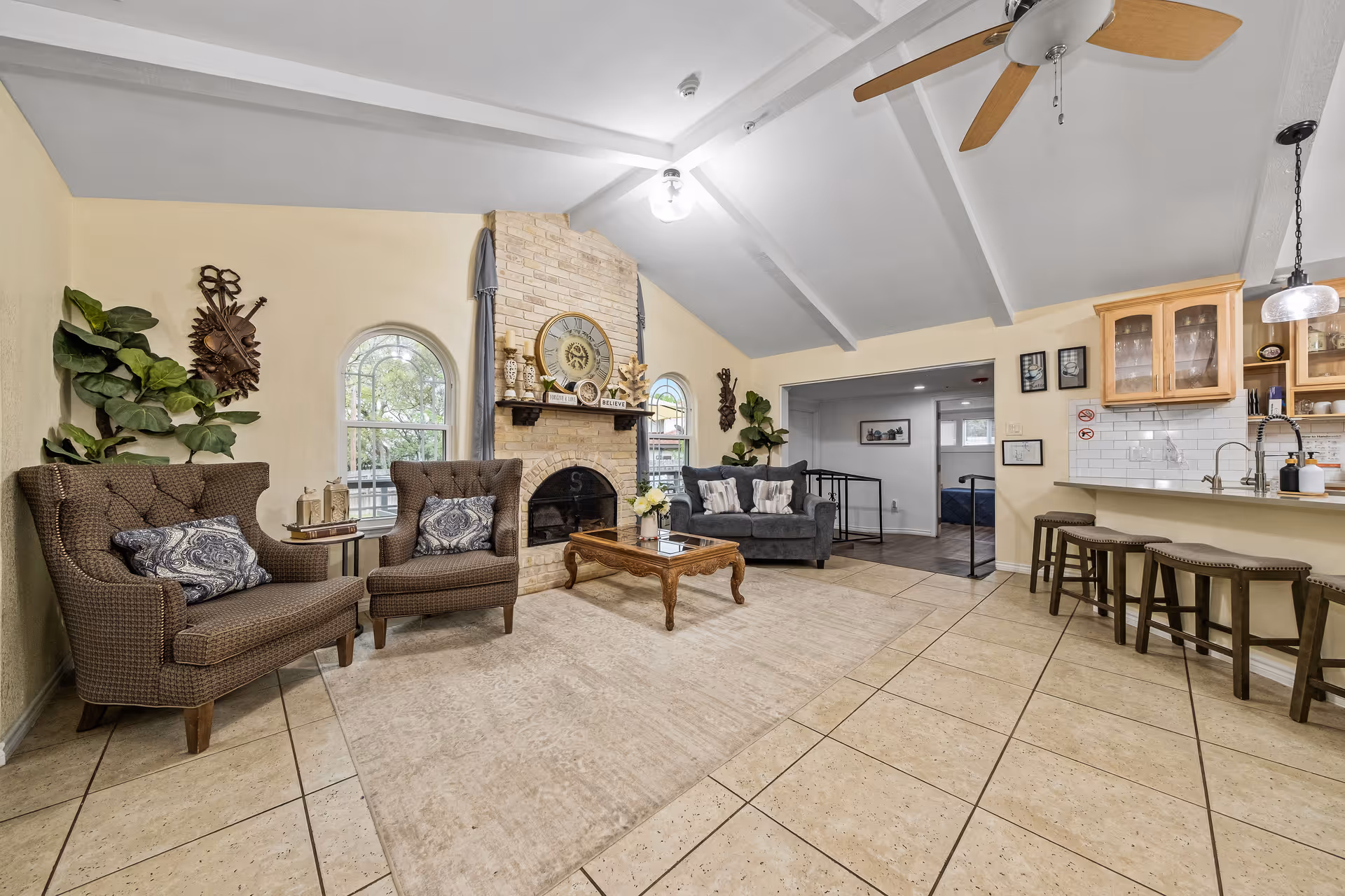 A cozy living room area with a high vaulted ceiling and ceiling fan. The room features a brick fireplace with decorative items on the mantel, two brown armchairs with patterned cushions, a wooden coffee table, and a gray sofa with pillows. There are two arched windows with curtains, a large indoor plant, and a tiled floor with a large area rug. To the right is a kitchen counter with four bar stools and wooden cabinets with glass doors.