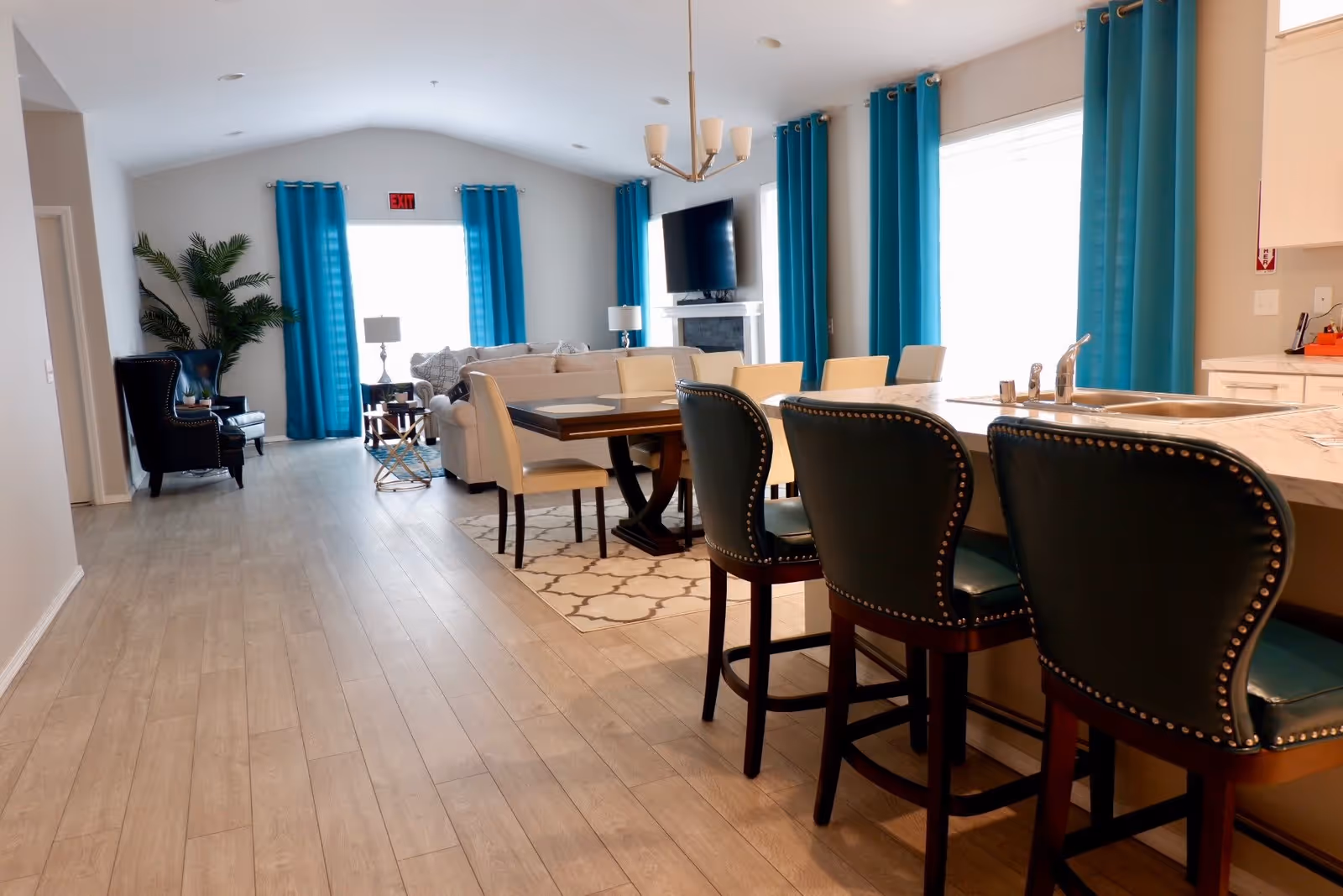 A bright and spacious interior of Mount Zion Assisted Living Home featuring a modern open-plan living area. The room includes a kitchen island with three dark green cushioned bar stools, a dining table with beige chairs, a cozy living room with beige sofas, a wall-mounted TV above a fireplace, and large windows with vibrant blue curtains. Light wood flooring and neutral walls create a warm and inviting atmosphere.