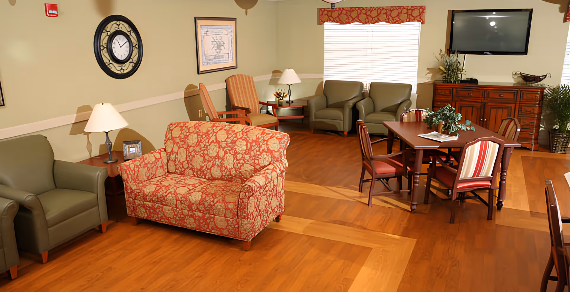 Community lounge with a patterned loveseat, armchairs, a dining table, TV and hardwood floors.
