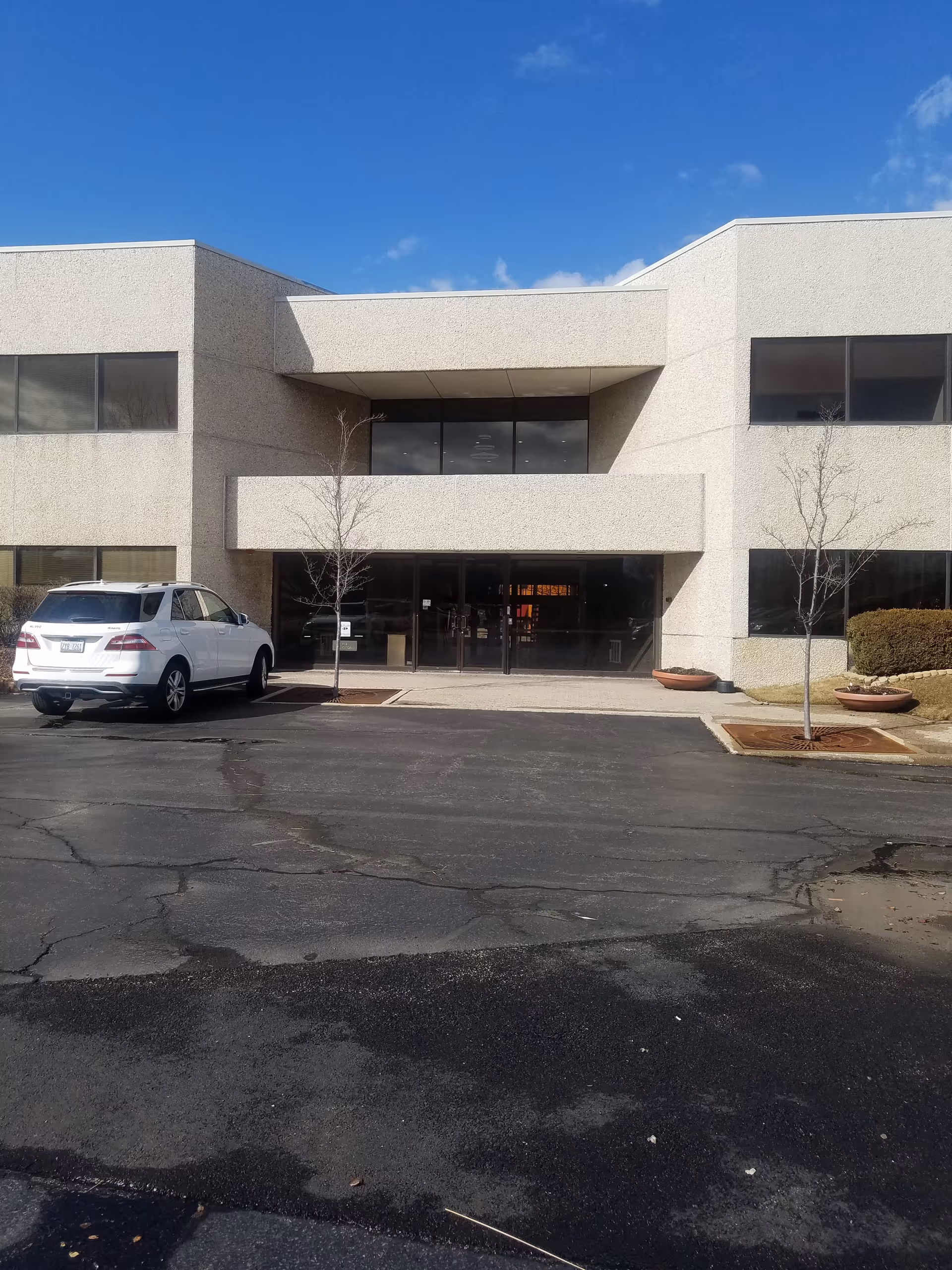 Two-story beige building with glass entrance and a parking lot in front.