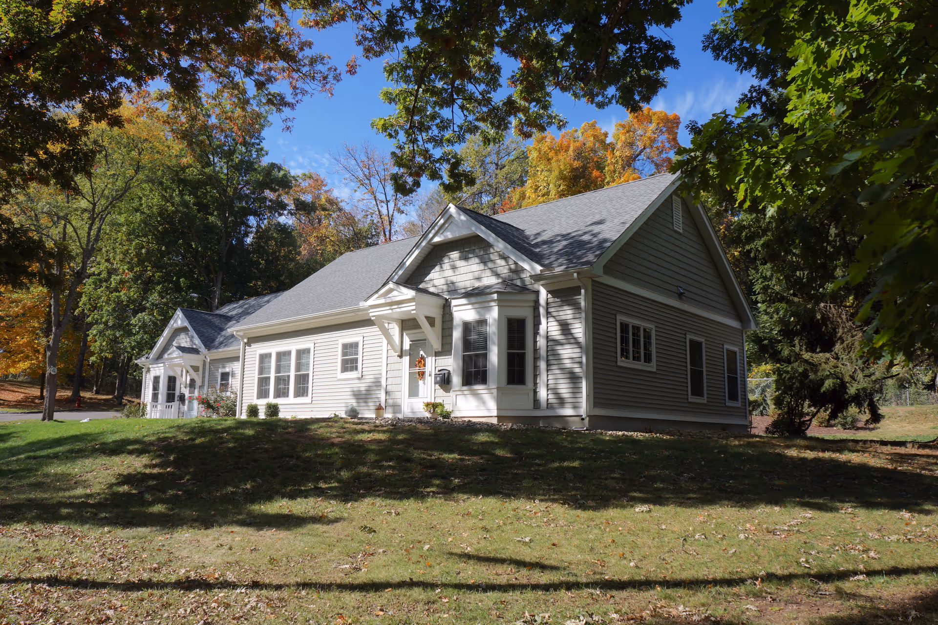 Light-gray single-story residential building with a small porch and multiple windows on a grassy, tree-lined lot in autumn.