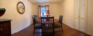 A dining area with a round wooden table surrounded by four dark wooden chairs. The room has light-colored walls, a wooden floor, a round mirror on the left wall, a window with floral curtains in the center, and a closet with louvered doors on the right.