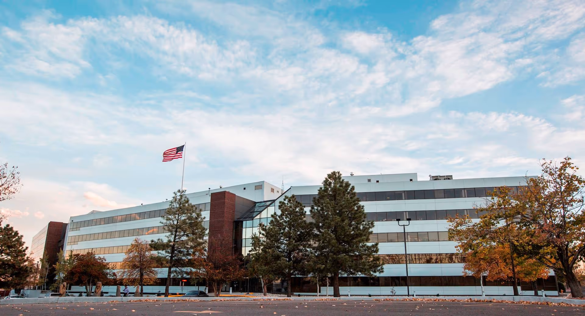 A large modern building exterior with an American flag, trees, and a parking lot.