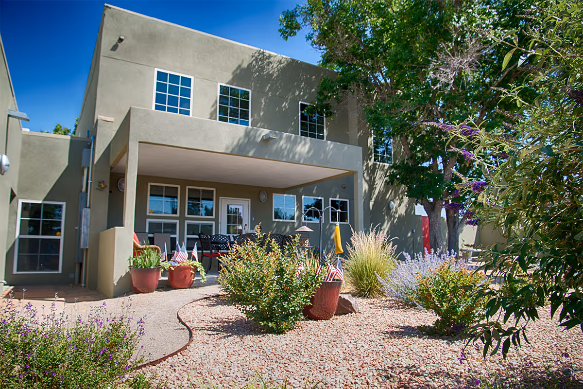 Exterior view of a two-story building with a covered patio area furnished with chairs and tables. The surrounding landscape features gravel ground cover, potted plants, bushes, and trees under a clear blue sky.
