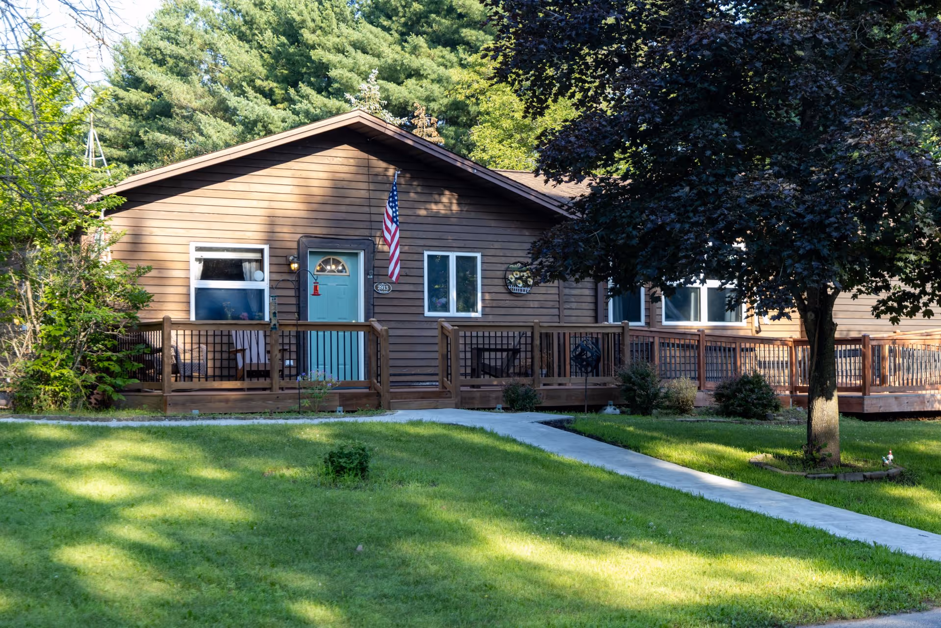Brown, single-story home with a wooden deck, flag, and green lawn.