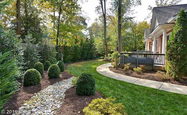 Well-kept outdoor garden with curved path, shrubs, trees, and a porch near a senior living building.