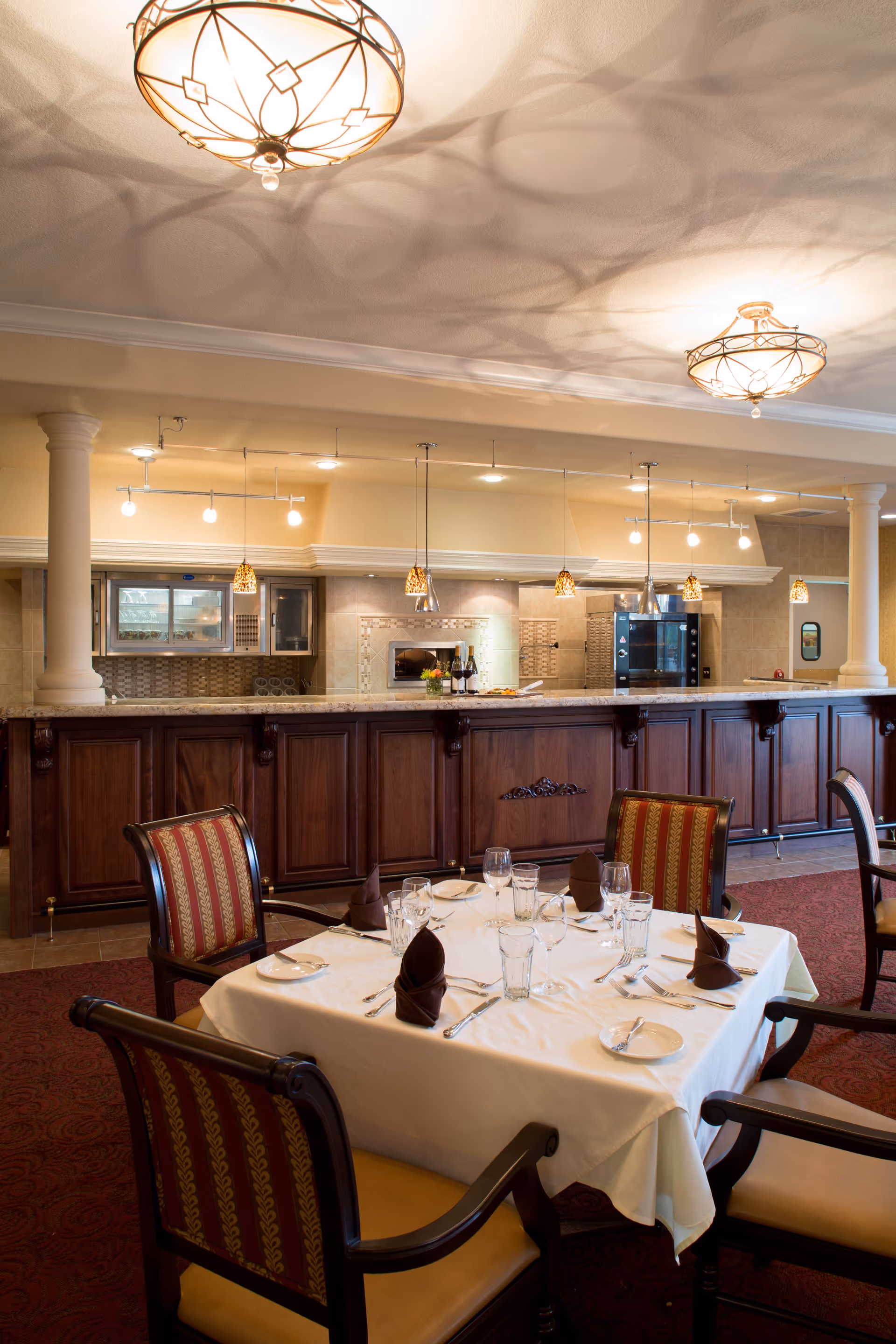 A dining area in a senior living facility with a table set for four people. The table is covered with a white tablecloth and has brown folded napkins, plates, glasses, and silverware. Behind the table is a long wooden counter with a kitchen area visible in the background, featuring pendant lights, a microwave, and an oven. The room has warm lighting and elegant ceiling fixtures.
