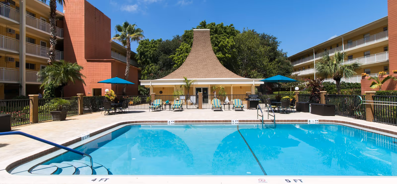 Outdoor swimming pool area at Scottish Towers II Apartments with lounge chairs, tables with umbrellas, palm trees, and a building with a unique sloped roof in the background under a clear blue sky.