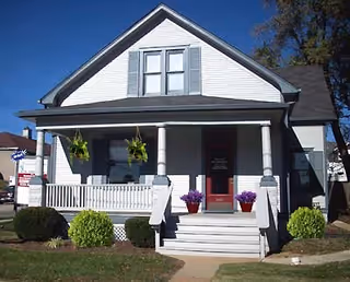 White, front-porch house with blue shutters, hanging plants, and potted purple flowers.