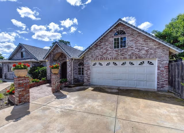 Brick single-story house front with a two-car garage, brick pillars topped with flower pots and a concrete driveway under a blue sky.