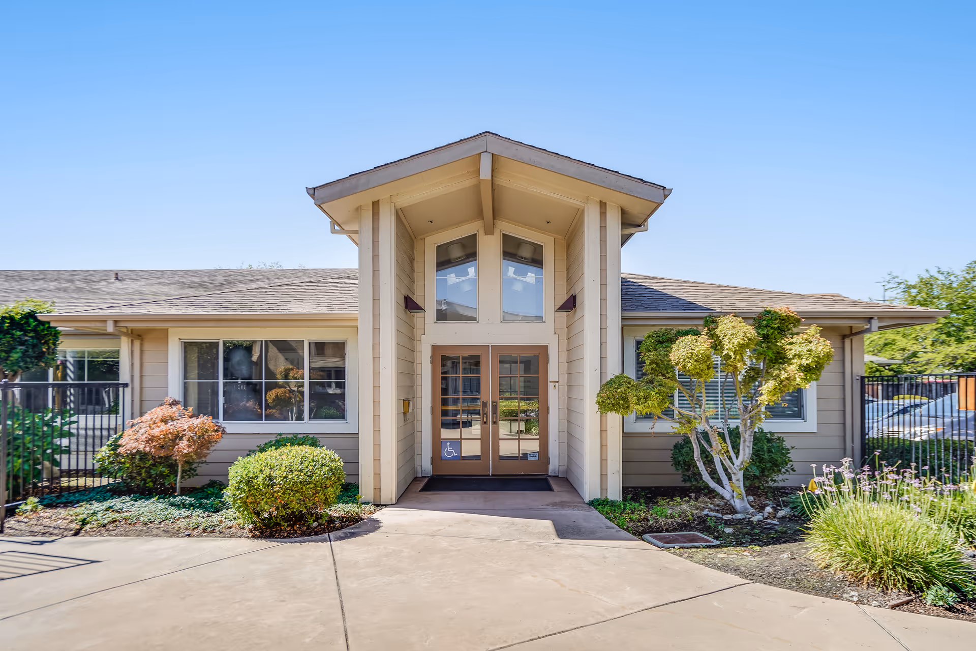 Front entrance of a single-story building with beige siding, large windows, and a peaked roof. The entrance has double glass doors with a wheelchair accessibility sign. There are neatly trimmed bushes and plants on either side of the walkway leading to the door under a clear blue sky.