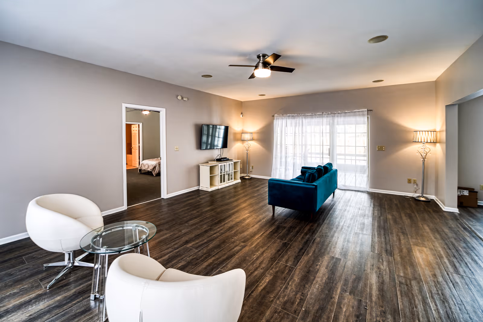 Spacious living room with teal sofa, white chairs, TV, and large windowed backdrop.