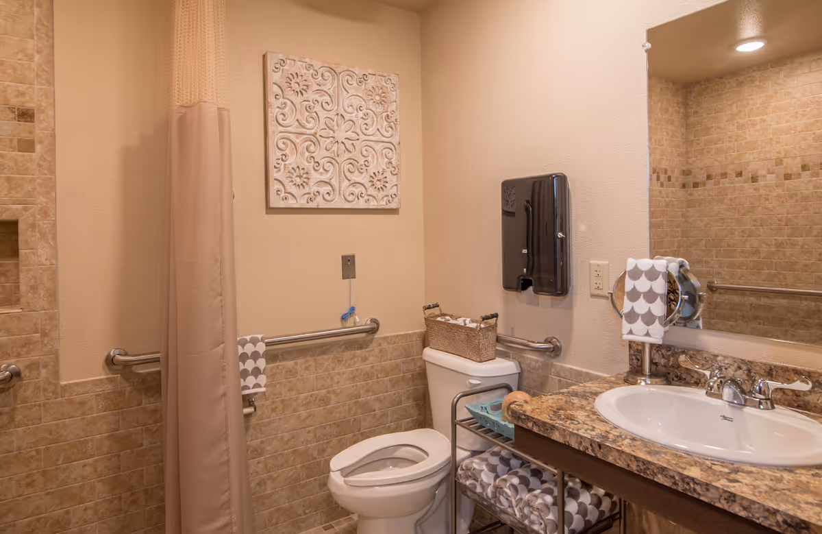 A clean and well-lit bathroom featuring a toilet with a basket on top, a sink with a granite countertop, a large mirror, and a shower area with a beige curtain. The walls are tiled halfway up with beige tiles and have grab bars installed for accessibility. A decorative wall art piece hangs above the toilet, and folded towels are neatly placed on a metal rack under the sink.