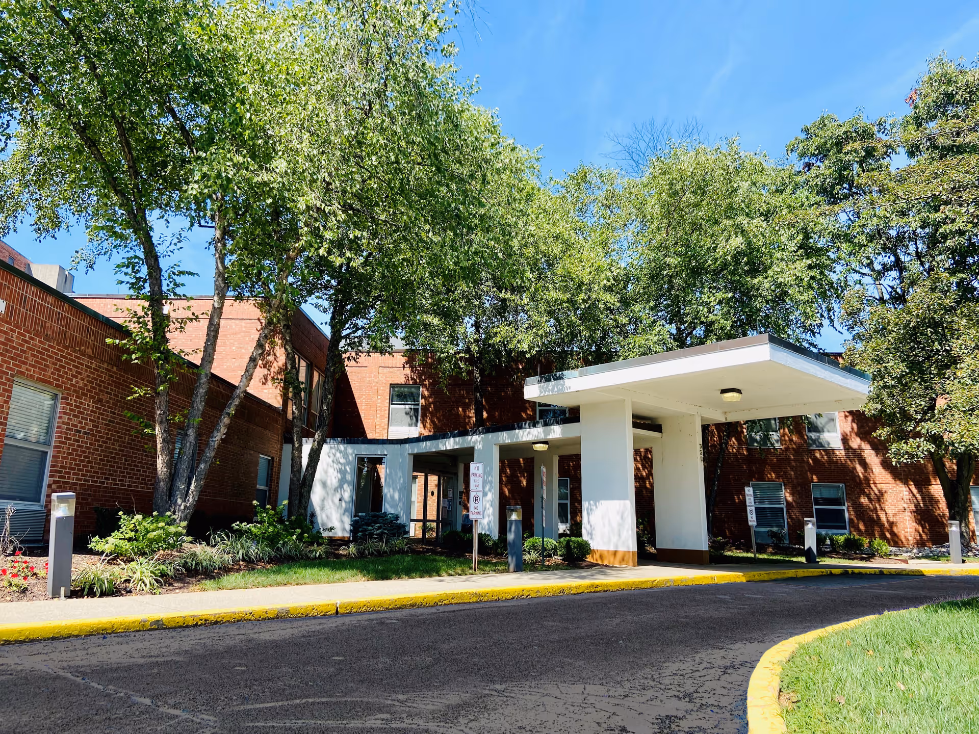 Front entrance of a brick building with a white covered drop-off canopy, trees, and landscaped beds.