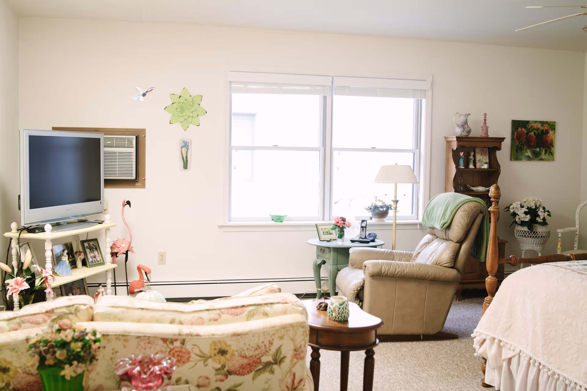 A cozy senior living room and bedroom area with a floral patterned sofa, a beige recliner chair, a wooden side table, and a bed with a light pink quilt. The room has a TV on a white stand, a window with blinds, various decorative items including flowers, framed photos, and wall art.