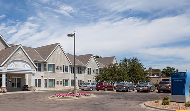 Two-story beige senior living building with a curved entrance, parking lot, and blue sign.