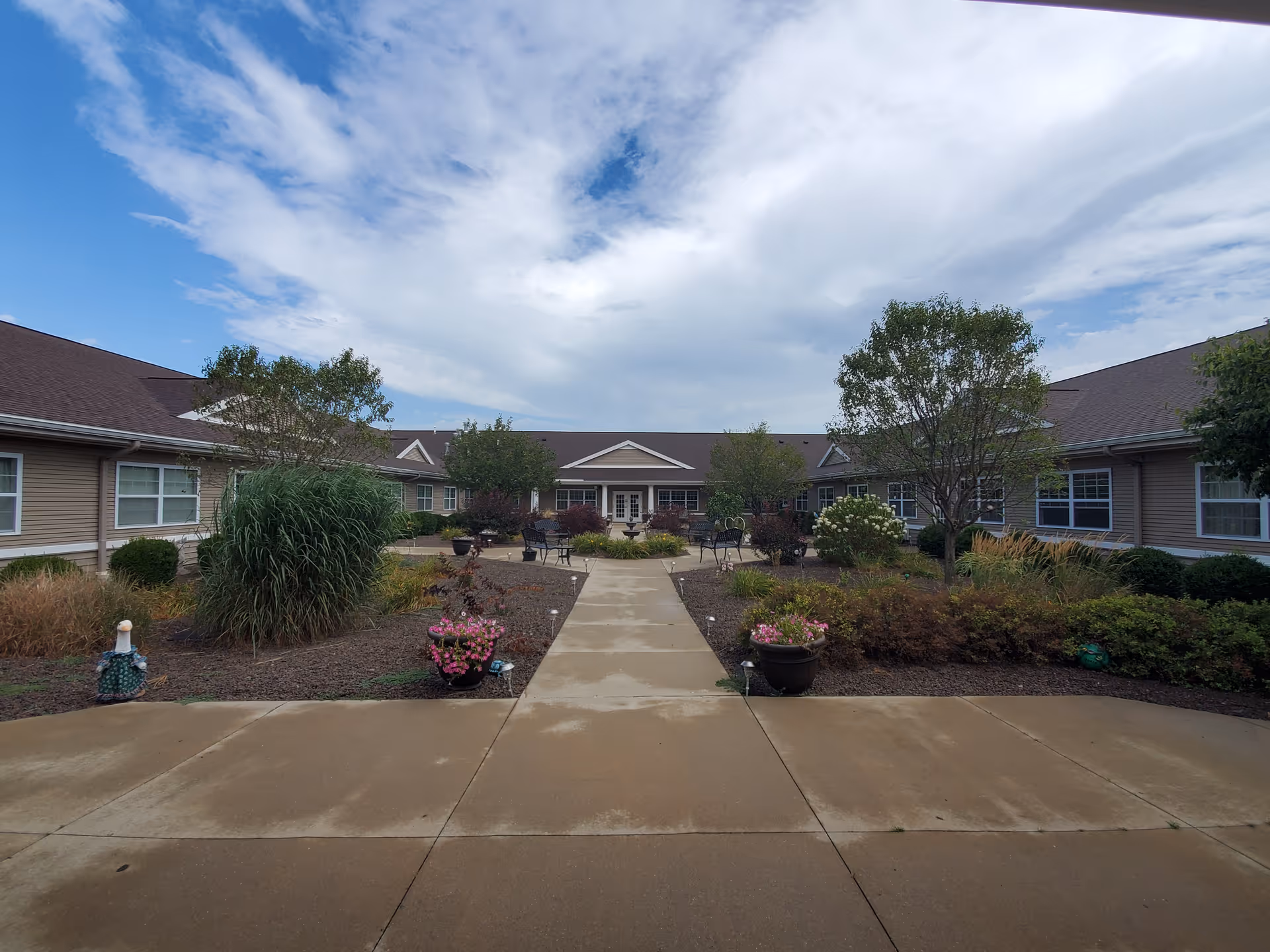 Outdoor courtyard area of The Pointe at Pontiac senior living facility with a paved walkway, landscaped garden beds with various plants and flowers, benches, and a partly cloudy sky overhead.